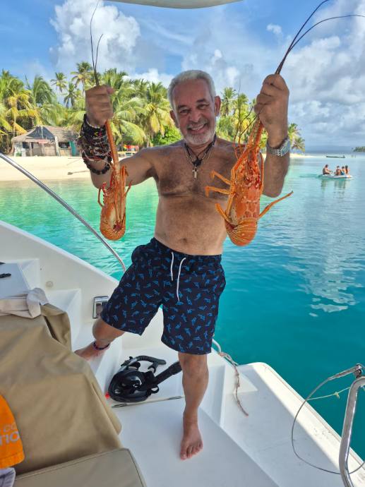 A guest holding two freshly caught spiny lobsters on the catamaran with a palm island behind