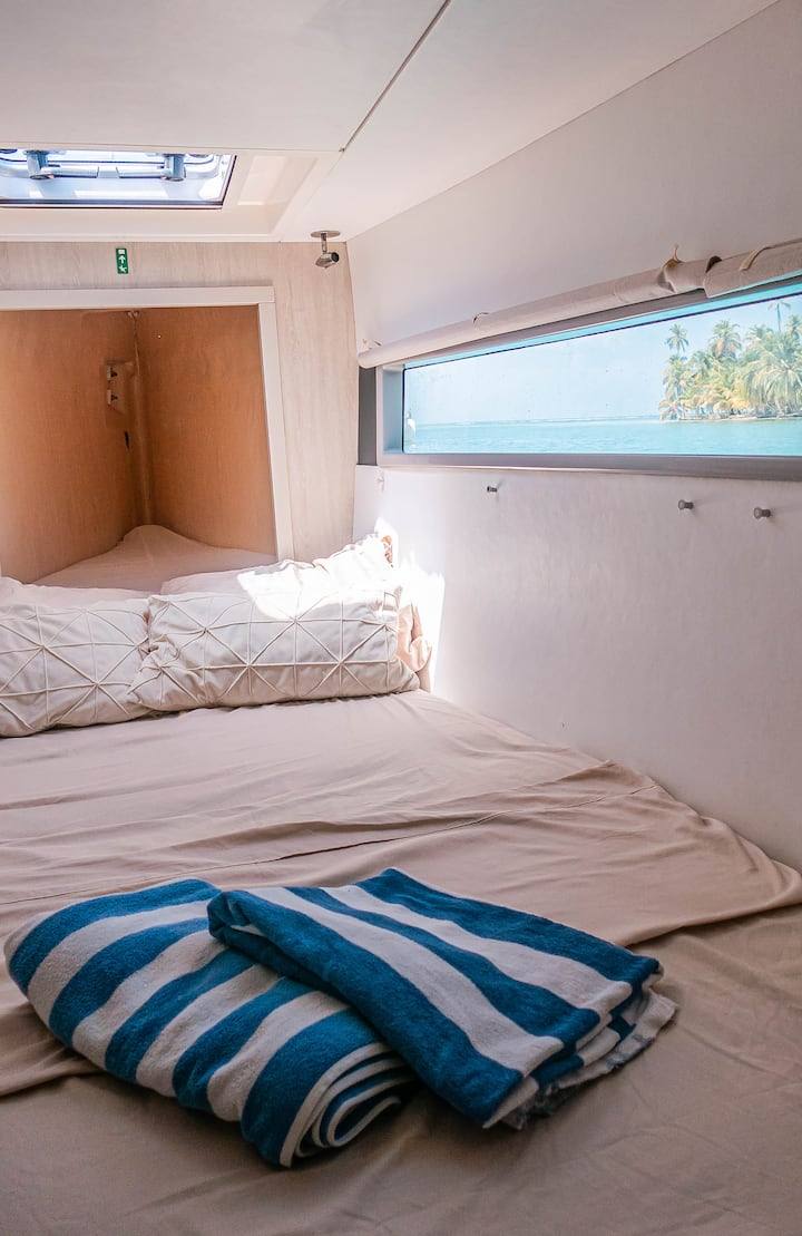 A forward double cabin with crisp linens, a skylight hatch and a sea-view window framing palm islands