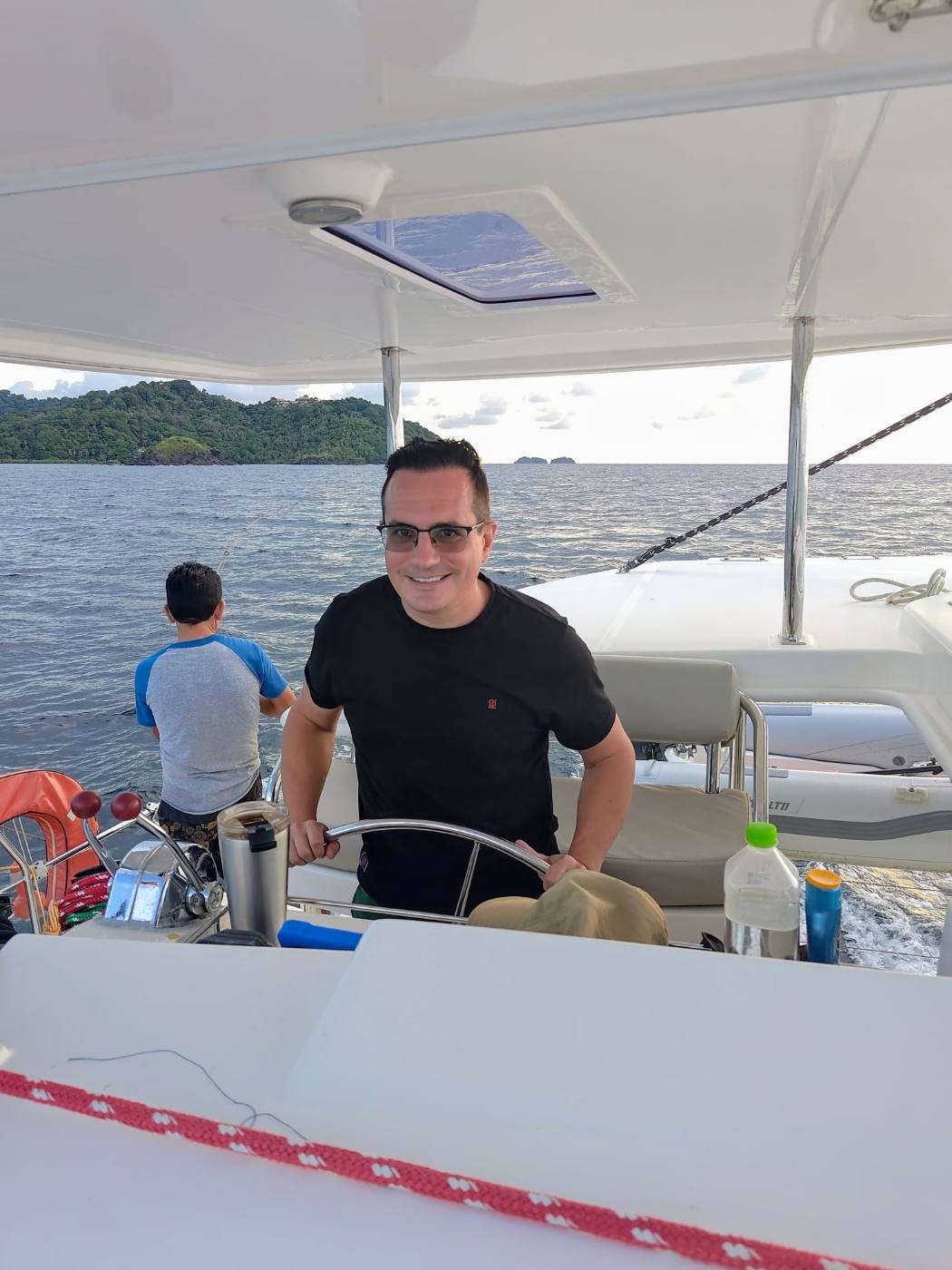 A smiling crew member giving a thumbs up from the deck, palm island and Breton flag behind