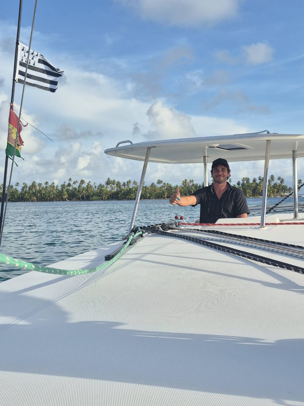 The captain smiling at the helm of the catamaran with a crew member fishing off the stern